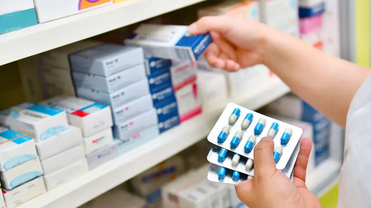 Ręce trzymają opakowania z kapsułkami, prawdopodobnie 3 dniowy antybiotyk na zapalenie ucha, na tle aptecznych półek z lekami.