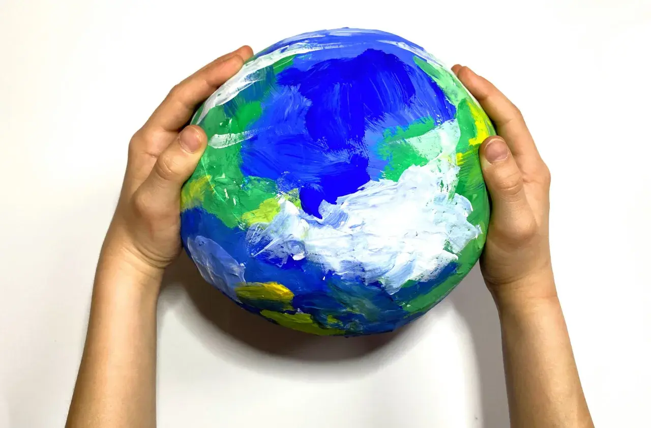 Dziecięca praca plastyczna przedstawiająca planetę Ziemię, namalowana farbami. Dłonie trzymają ją delikatnie, jakby chroniły nasz dom.