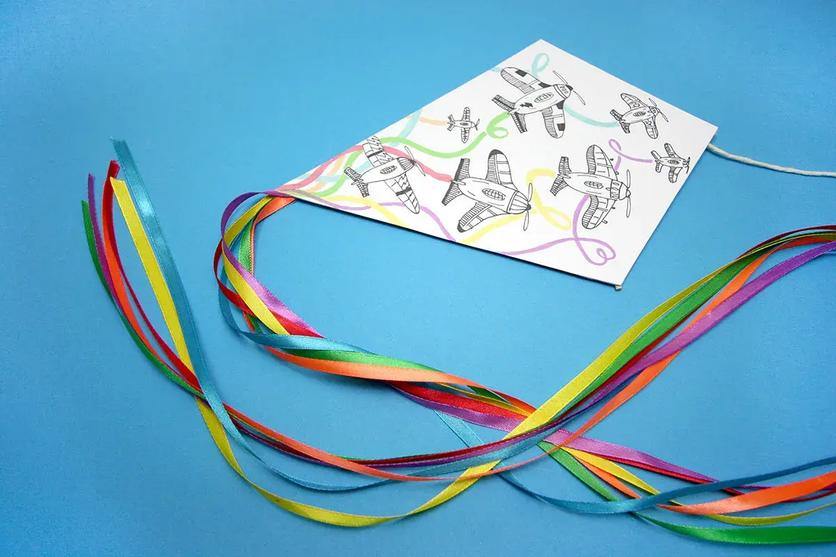 Kolorowy latawiec DIY z samolotami na tle niebieskim. Wstążki w tęczowych barwach tworzą ogon.