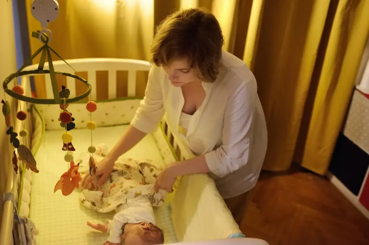 Mama uspokaja dziecko w łóżeczku, by złagodzić lęk separacyjny u niemowląt. Nad łóżeczkiem wisi karuzela z leśnymi zwierzątkami.