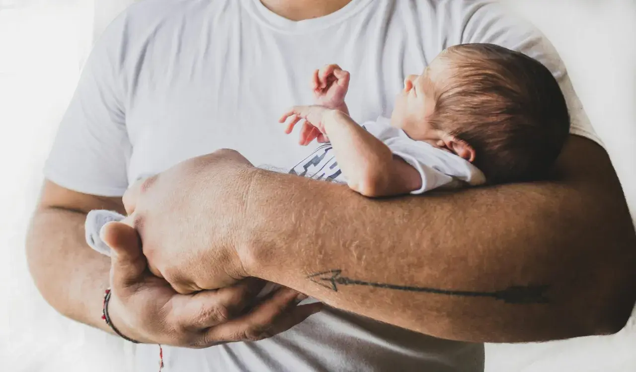 Ojciec tuli śpiącego niemowlaka. Maluch ma zamknięte oczy, a jego rączki delikatnie unoszą się w powietrzu. Tata z tatuażem strzały na przedramieniu czule obejmuje swoje dziecko.