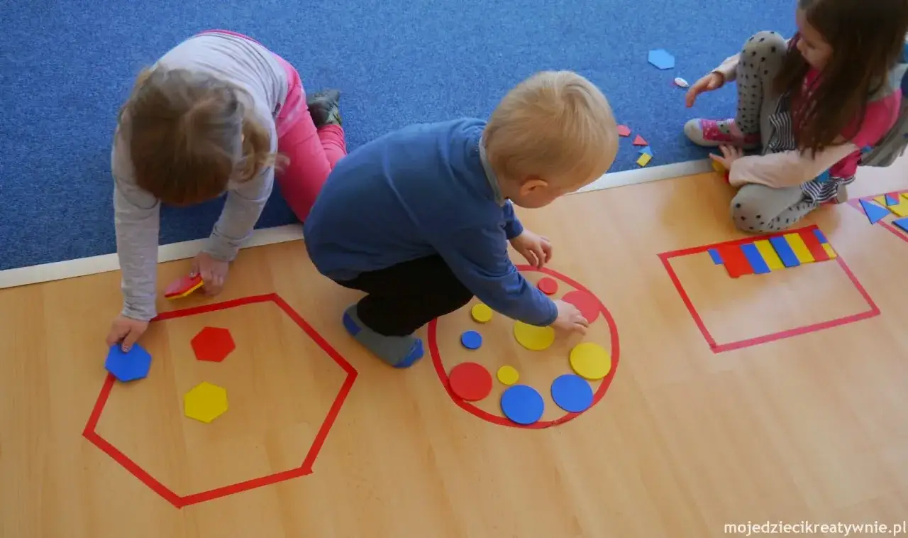 Dzieci w przedszkolu bawią się kolorowymi figurami geometrycznymi, tworząc wzory w wyznaczonych obszarach.