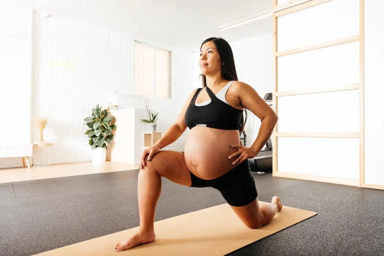 Kobieta w zaawansowanej ciąży wykonuje ćwiczenia w ciąży, rozciągając nogi na macie w jasnym pomieszczeniu.