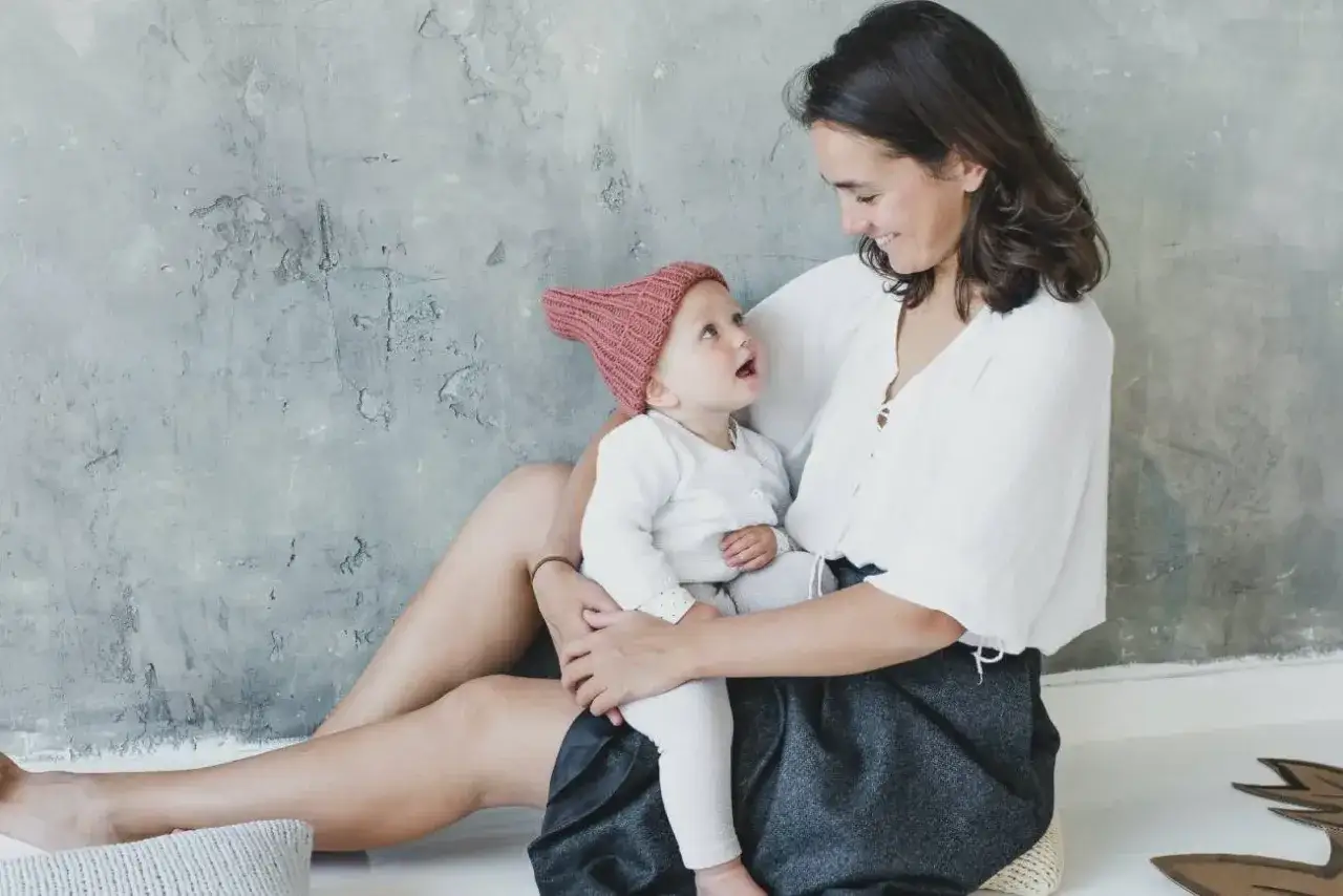 Uśmiechnięta mama trzyma na kolanach 6 tygodniowe dziecko w r&oacute;żowej czapeczce.