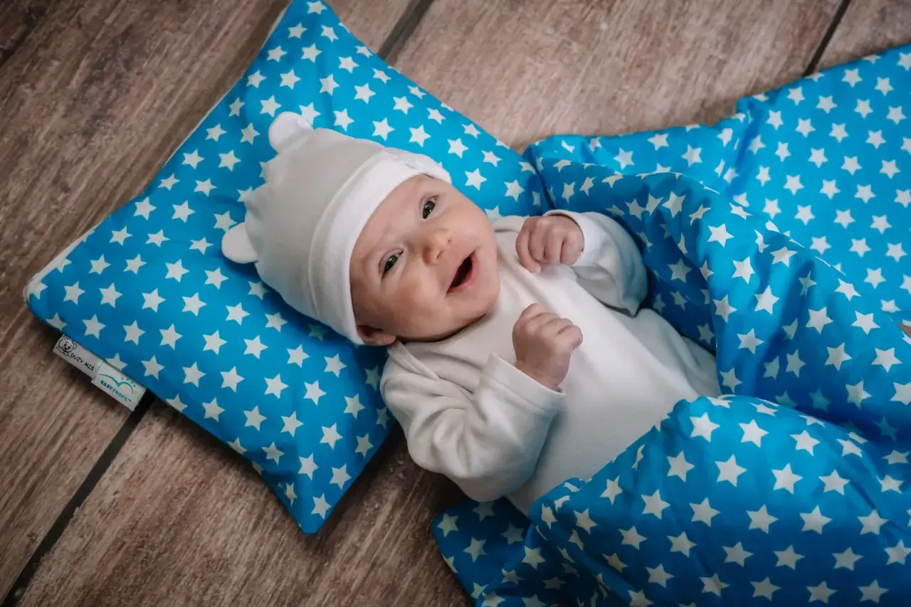 Uśmiechnięte dziecko w białej czapeczce i body, otulone niebieskim kocykiem w gwiazdki. Jak ubrać dziecko do snu bez śpiworku?