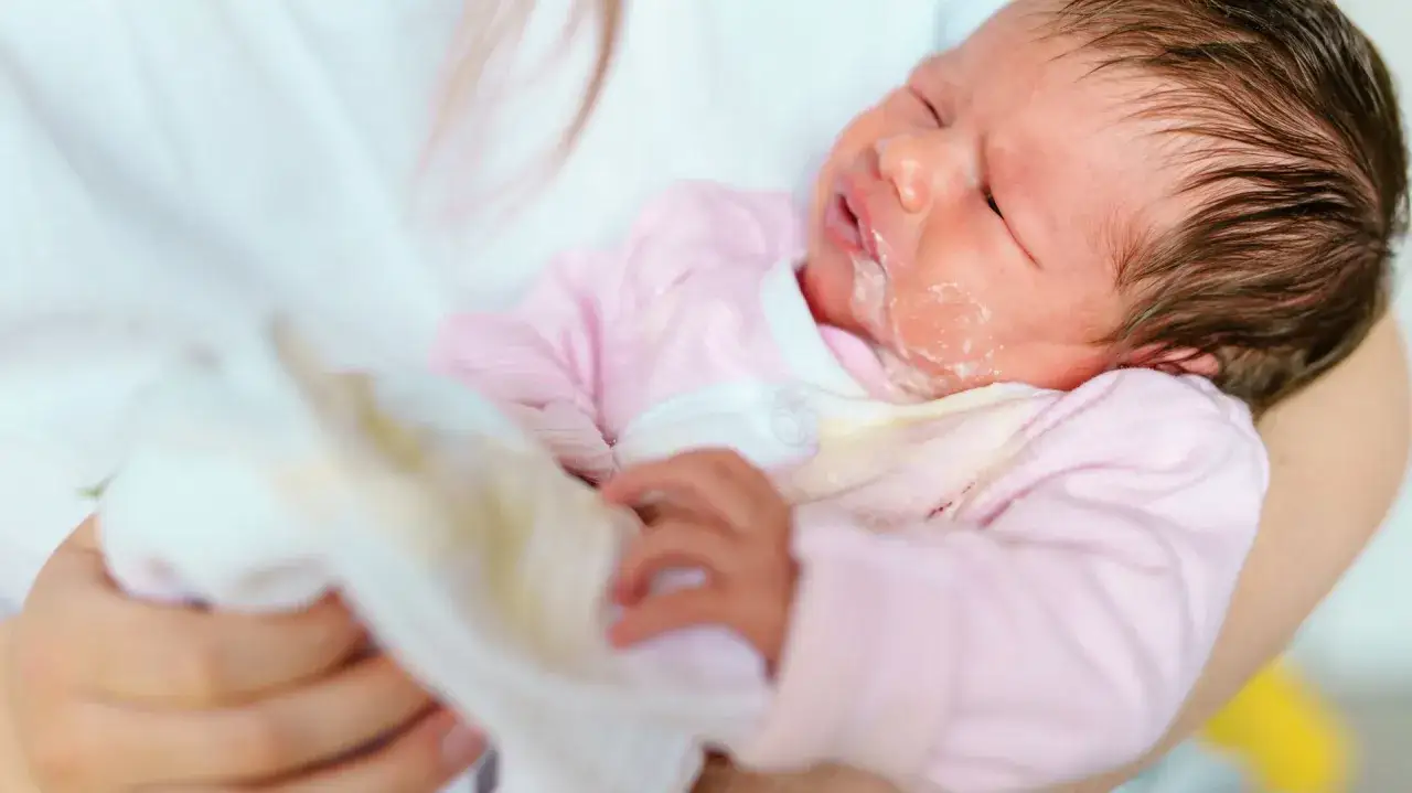 Noworodek w r&oacute;żowym pajacyku z widocznym ulewaniem. Mama wyciera buźkę dziecka.