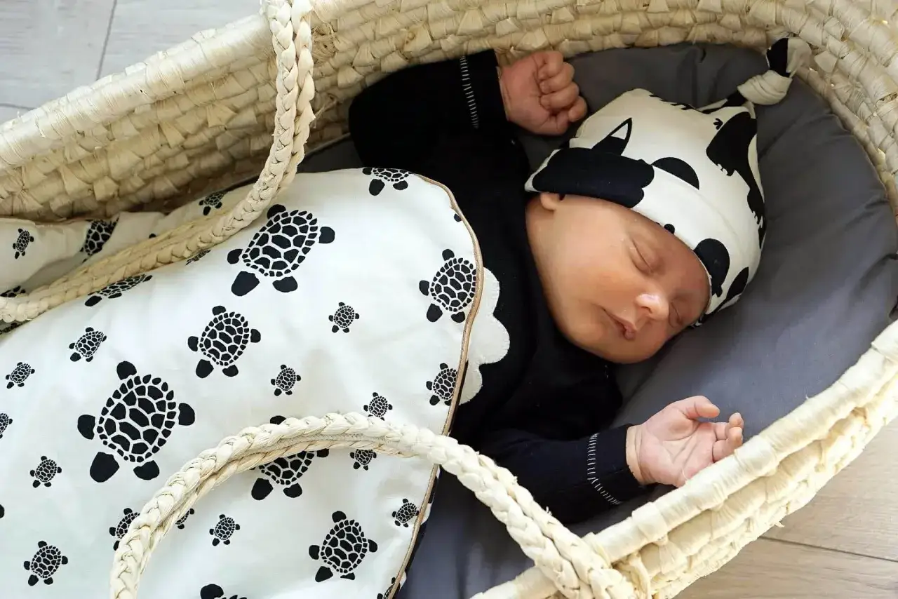 Niemowlę śpi w koszu, otulone kocykiem z żółwiami. Czarna czapeczka z tym samym motywem dopełnia stylizację, pokazując, jak ubrać niemowlę do snu.