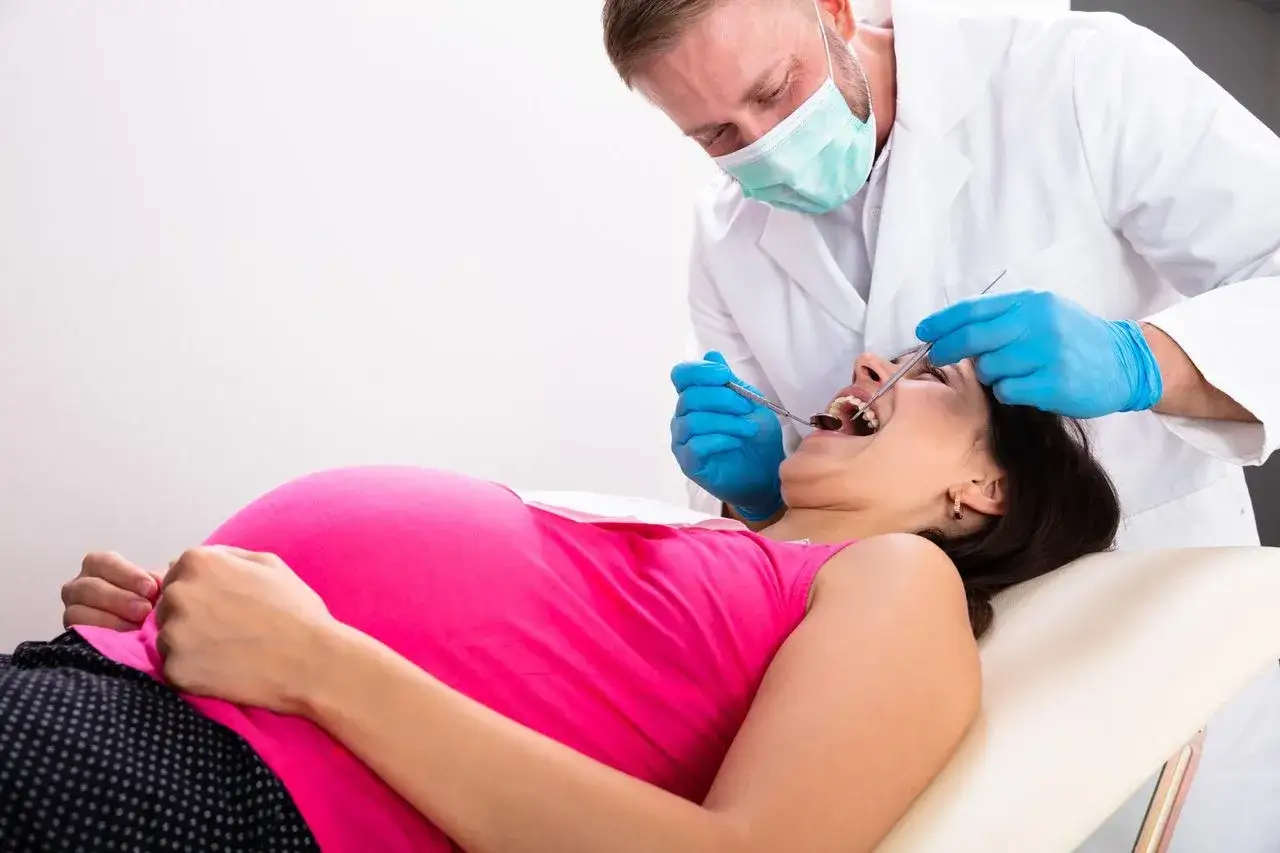 Dentysta w ciąży bada zęby pacjentki. Kobieta leży na fotelu, a lekarz w rękawiczkach i masce wykonuje badanie.